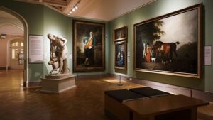 A well-lit art gallery at the Appleton Museum of Art displays several large, framed classical paintings on green walls. A white marble statue of a man with a dog stands prominently on the left. Wooden benches are positioned in the center of the room, and a wooden floor contrasts with the artwork.