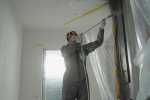 A person wearing protective gear expertly uses a spray paint gun on a wall in a room marked for rehab to sell. The space is covered with plastic sheeting for protection, and bright light filters through a window in the background. The individual dons goggles and a mask for safety.
