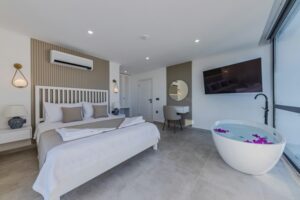 Modern, minimalist bedroom with a large bed, wall-mounted TV, and a freestanding bathtub filled with water and flower petals. Perfect for staging a home, the room features light grey tiles, a round mirror above a desk, wall-mounted lamps, and a large window allowing natural light to enter.