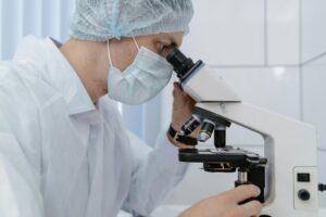 A person wearing a white lab coat, face mask, and hair cover is carefully examining coronavirus samples through the eyepiece of a microscope in a laboratory.