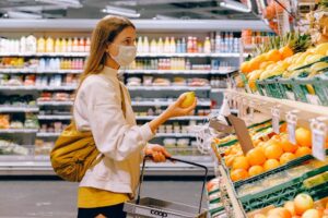 During the pandemic, a person wearing a face mask and a yellow backpack is holding an apple while shopping for produce in a grocery store. They have a small shopping basket in their other hand. Shelves stocked with various fruits, vegetables, and packaged items are visible in the background.