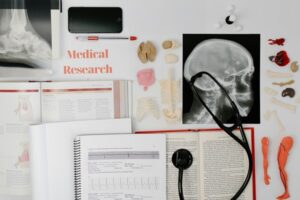 A cluttered workspace featuring medical research materials, including textbooks, anatomical models, an X-ray of a skull, a stethoscope, a smartphone displaying COVID-19 data, a pen, and a notebook with graphs. Text reads "Medical Research.