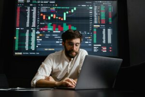 A man with glasses and a beard in a white shirt works on a laptop at a desk. Behind him, a large screen displays stock market data, reflecting the world economy through charts, graphs, and figures. The environment is dark, and the screen's data is well-lit.