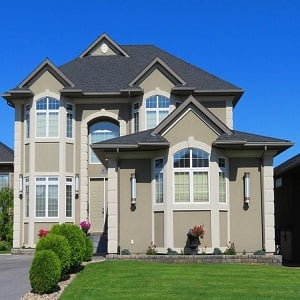 A charming two-story beige house with dark gray roofing stands under a clear blue sky, perfect for those immersed in home searches. It boasts arched windows and small shrubs in the front yard, complemented by a neatly trimmed green lawn.