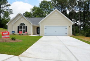A beige, single-story house with a dark gray roof and a two-car garage is shown. The front yard features well-maintained grass and a concrete driveway. Signs in the yard advertise "Private" and "Backyards." Trees are visible in the background, highlighting its appeal in The Ocala sellers market.