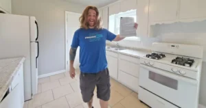 A person with long hair, wearing a blue shirt and gray shorts, stands in a kitchen holding up a piece of paper. The kitchen has white cabinets, a white gas stove, and a refrigerator. The person appears to be smiling and excited—perhaps they've just discovered an exciting Ocala real estate investment opportunity!