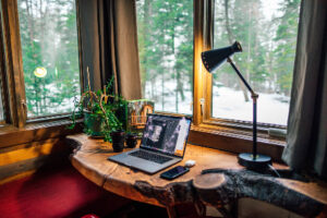 A cozy workspace setup featuring a wooden desk by a window overlooking a snowy forest. On the desk, there's an open laptop, a smartphone, a desk lamp, and some potted plants. Natural light streams in, creating a warm and inviting atmosphere that epitomizes Ocala Home Decor charm.