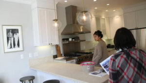 Two people are in a modern kitchen. One person stands near a stainless steel stove and hood, facing towards a countertop. The other, wearing a plaid shirt, is holding a book or magazine about how to sell my Ocala house. The kitchen has white cabinets, pendant lights, and a framed black-and-white photo on the wall.