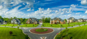 A panoramic view of a suburban neighborhood featuring a circular roundabout surrounded by large, brick and modern-style houses. Lush green lawns and trees line the streets under a bright blue sky with scattered clouds, creating an ideal setting if you're looking to sell my Ocala house.