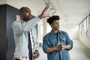 Two individuals are standing indoors at the Ocala open house. The person on the left, wearing a light jacket, is gesturing as if explaining something. The person on the right, in a darker jacket, is looking towards where the first person is pointing. They seem to be in a discussion.