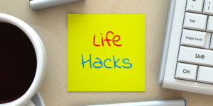 A bright yellow sticky note with "Life Hacks" written in red and blue marker is placed on a wooden desk. A cup of black coffee, part of a white keyboard, and the end of a pen can also be seen on the desk, hinting at a productive morning dive into the latest interest rate hack.