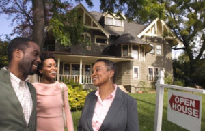 Three people stand together, smiling, in front of a large, two-story house with an "Open House" sign in the yard. The house is surrounded by greenery and trees under a clear sky. They seem to be enjoying the nice weather and discussing interest rates while appreciating the property.