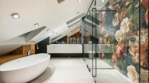 A modern bathroom with a white freestanding bathtub on the left, a glass-enclosed shower featuring a colorful floral wall on the right, and a white countertop with cabinets and mirrors in the background. The ceiling is sloped with recessed lighting that feels like an interest rate hack for your home's value.