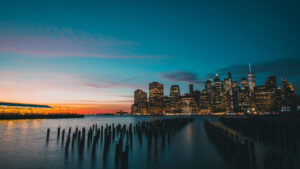 A twilight view of a city's skyline from across a body of water. The sky is a gradient of orange, pink, and blue, reflecting off the water's surface. Numerous wooden posts protrude from the water in the foreground, leading the eye toward the illuminated buildings—much like Ocala real estate surging in appeal.
