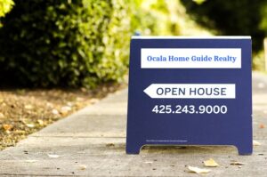 A sidewalk sign reads "Ocala Home Guide Realty" at the top, followed by "Ocala Open House," and a phone number: 425.243.9000. The sign is set on a concrete path with blurred greenery in the background.