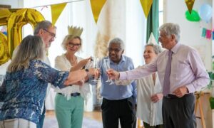 A group of older adults, wearing festive paper crowns, stand in a semi-circle and clink their glasses together in a celebratory toast. They appear cheerful and are surrounded by colorful decorations and balloons, enjoying the event’s free services.