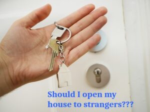 A hand holding a keychain with keys and a house-shaped charm in front of a door lock. The text overlay at the bottom in blue reads, "Should I open my Ocala house to strangers???