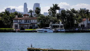 A luxurious waterfront house bordered by palm trees with a white yacht moored at the dock. The skyline of a city with tall buildings is visible in the background, across the blue water. Amidst Ocala real estate surging, this scene exudes a tropical and affluent ambiance.