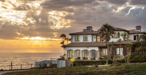 A Mediterranean-style house with clay roof tiles and large windows sits by the ocean under a dramatic sky filled with clouds and a setting sun. The golden hues of the sunset reflect on the water and sky, casting a warm glow over the scene. Palm trees surround this gem as Ocala real estate surges.