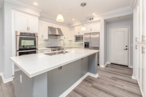 A modern kitchen with white cabinets, stainless steel appliances, and a large island in the center becomes a focal point in home searches. The island features a built-in sink and sleek white countertop. Pendant lights hang above, while light gray walls and light wood flooring complete the look.
