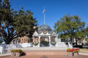 A decorative gazebo with a flagpole on top is nestled among large trees in a park, reminiscent of the charm seen in SE Ocala houses for sale. The gazebo has a dome-shaped roof and is adorned with festive holiday wreaths. Several large flower pots with red and yellow blossoms are placed in front.