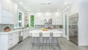 A modern kitchen with light gray cabinets, a large marble island featuring four white chairs, and a stainless steel backsplash. Three pendant lights illuminate the space, while a window bathes it in natural light. A fruit bowl sits atop the island—find similar designs at Ocala Homes Online; Photo by Zack Gudakov on Upsplash.