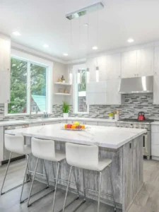 A modern kitchen with light gray cabinets, a large marble island featuring four white chairs, and a stainless steel backsplash. Three pendant lights illuminate the space, while a window bathes it in natural light. A fruit bowl sits atop the island—find similar designs at Ocala Homes Online; Photo by Zack Gudakov on Upsplash.
