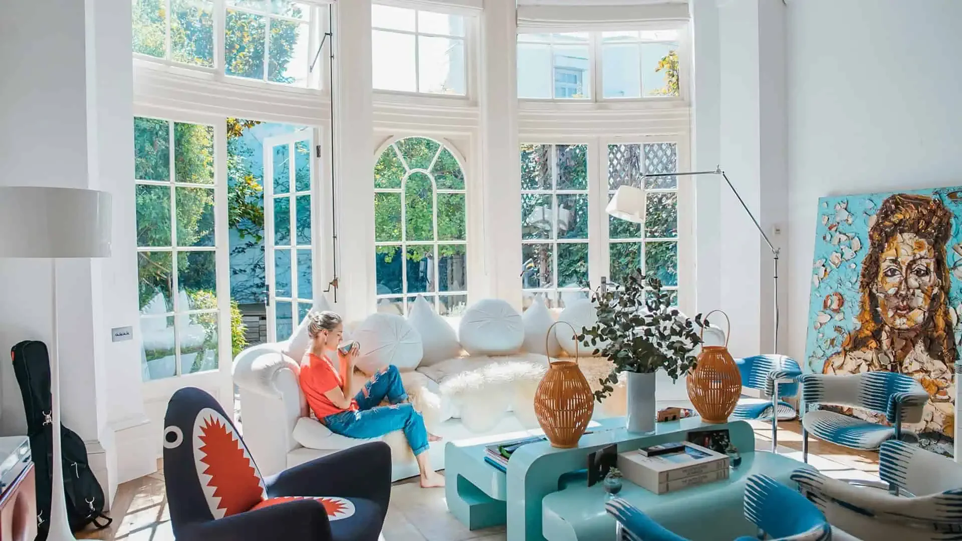 In this Ocala Homes Online background image, a woman in a red shirt sits on a white sofa in a bright living room with large windows. The room features modern furniture, including a teal table, wicker lanterns, and a colorful painting on the wall. Sunlight streams through the windows.